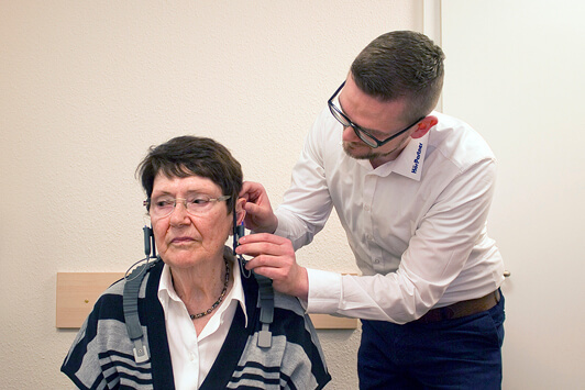 Ein Hörakustiker passt einer Seniorin in einer Hörgeräteberatung ein Hörgerät an.